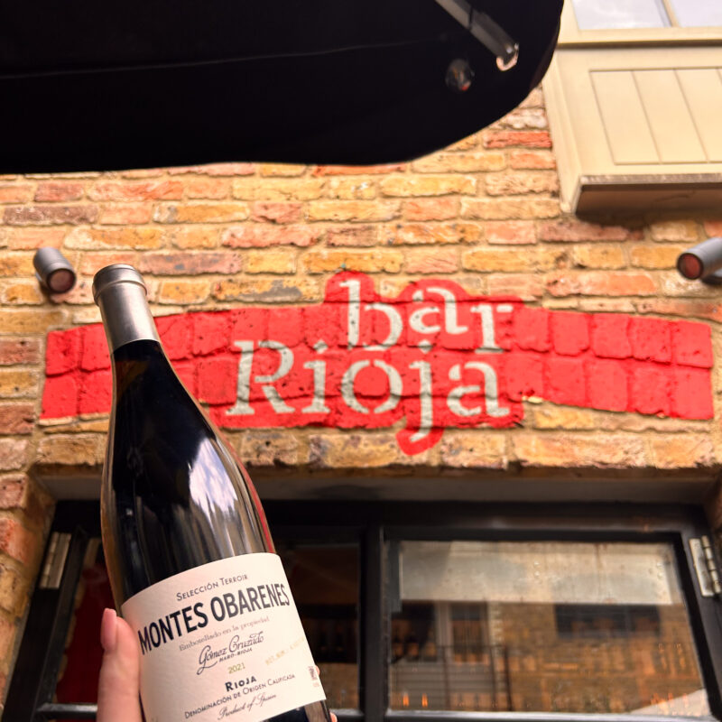 Bottle of white wine being held up in front of the door to Bar Rioja. The Bar Rioja logo is visible in the background